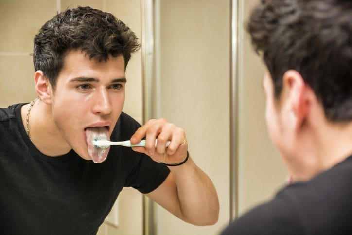 man brushing his tongue looking at a bathroom mirror